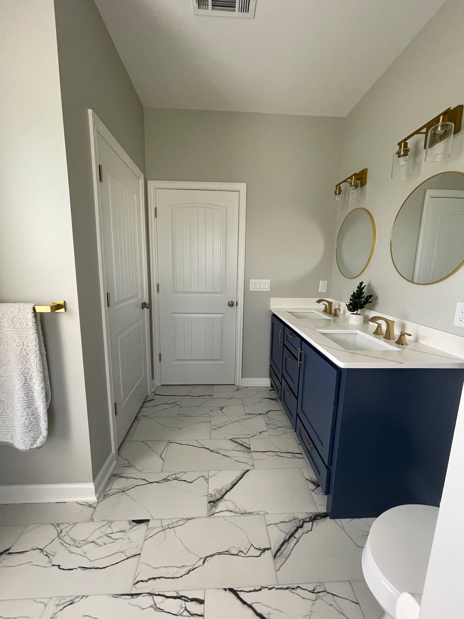 Texas Pro Painters: Luxurious bathroom featuring a navy blue double vanity with gold fixtures, marble-look floor tiles, light grey walls, and two round gold-framed mirrors, highlighting quality interior painting.