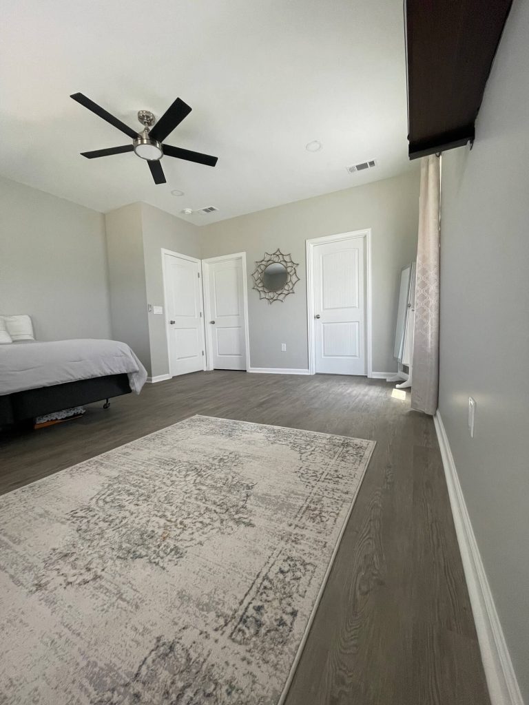 Texas Pro Painters: Modern bedroom with light grey walls, dark wood-look laminate flooring, white doors, a ceiling fan, and a light-colored area rug, showcasing fresh interior painting.