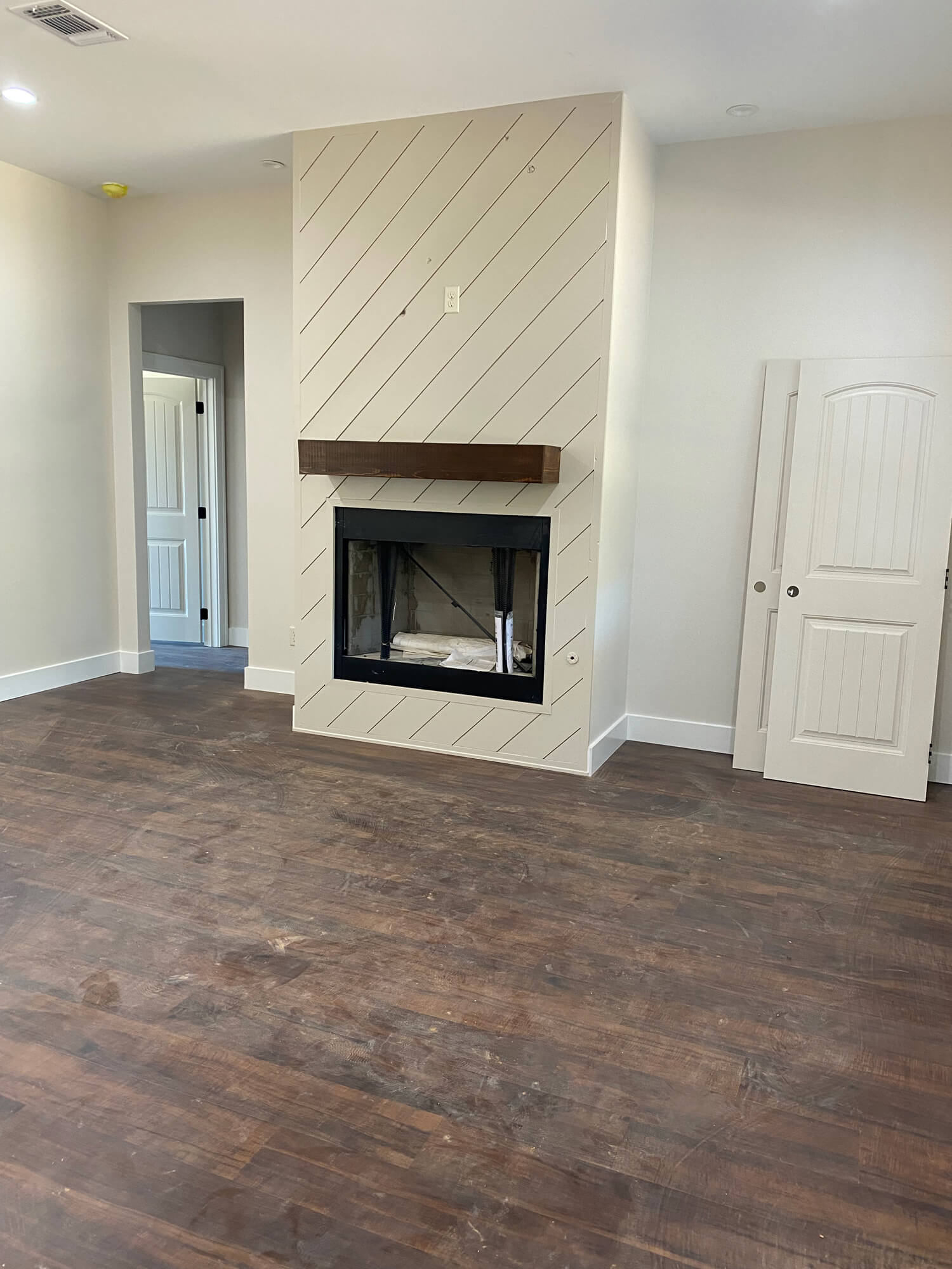 Texas Pro Painters: Unfurnished living room with a newly painted beige fireplace featuring diagonal shiplap and a dark wood mantel, light walls, and dark wood flooring, ready for final touches.