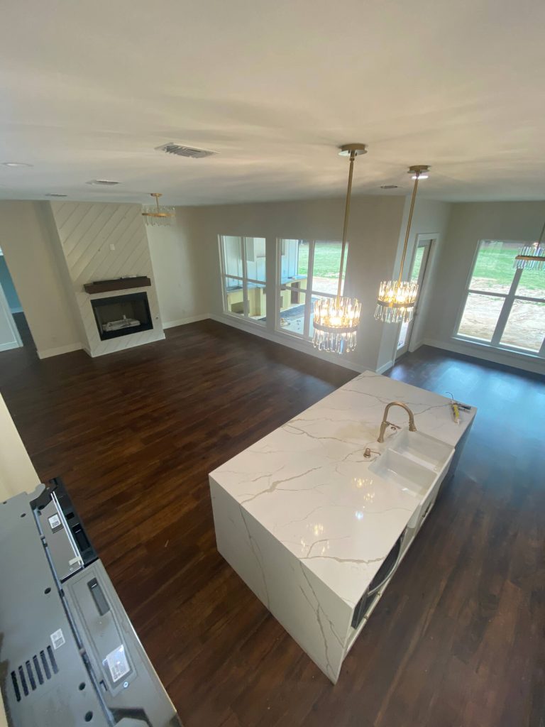 Texas Pro Painters: Open-concept living space with dark wood flooring, a white waterfall kitchen island, a modern fireplace, and large windows, showcasing fresh interior paint and finishes.