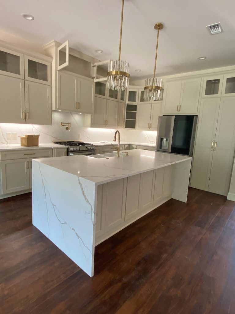 Texas Pro Painters: Newly installed high-end kitchen featuring light beige cabinets, a waterfall island with marble-look countertops, dark wood floors, and elegant gold light fixtures, perfect for cabinet painting and refinishing.