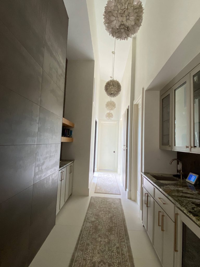 Texas Pro Painters: Luxurious hallway with a wet bar featuring light grey cabinets and granite countertops, modern light fixtures, a long runner rug, and a dark accent wall, showcasing sophisticated interior finishes.