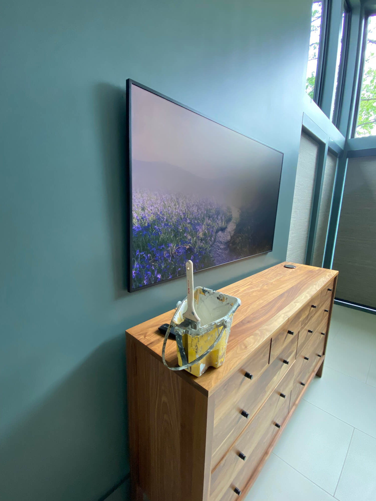 Texas Pro Painters: Interior room with a freshly painted teal accent wall, a wall-mounted TV displaying a nature scene, a wooden dresser, and painting supplies, showcasing professional interior painting.