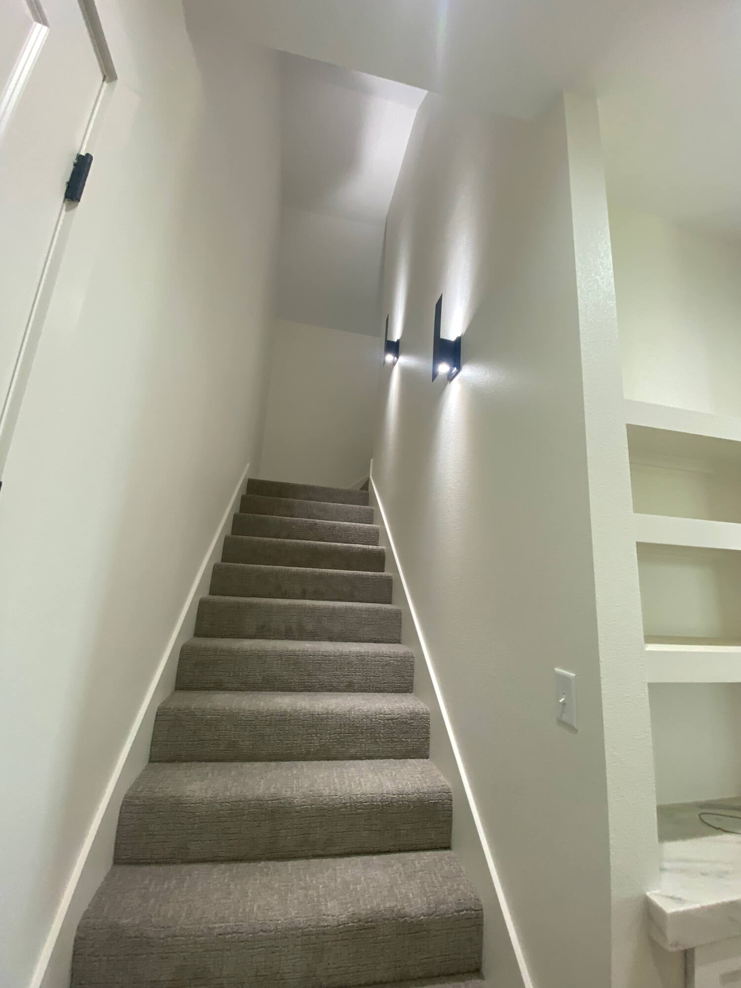 Texas Pro Painters: Modern staircase with light grey carpeted steps, freshly painted light beige walls, sleek wall-mounted lighting, and built-in shelving, demonstrating quality interior painting.