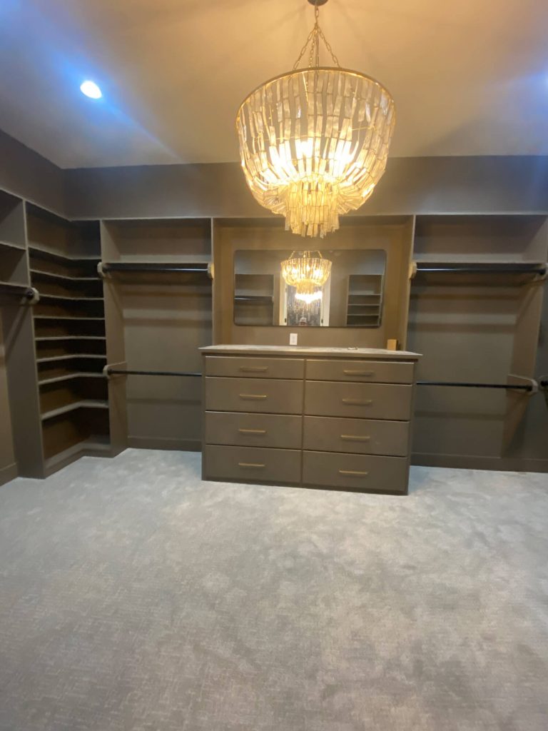Texas Pro Painters: Custom walk-in closet featuring built-in shelving, dark grey painted walls and cabinetry, light grey carpet, and an ornate crystal chandelier, showcasing specialized interior painting.