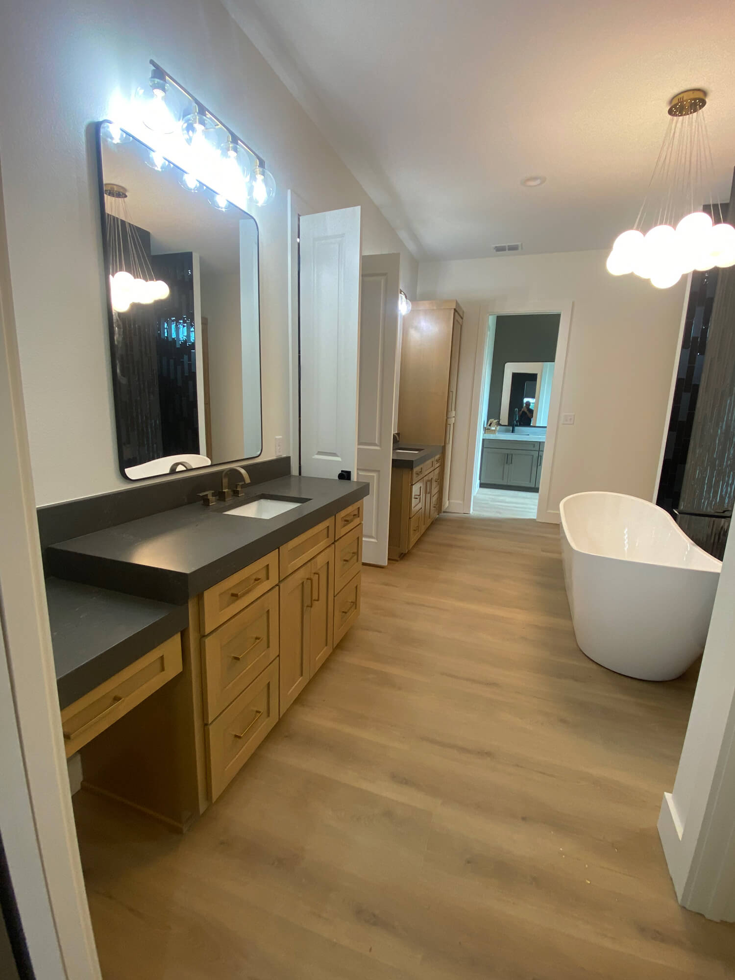 Texas Pro Painters: Luxurious modern bathroom featuring light wood vanities with dark countertops, a large free-standing tub, wood-look flooring, and unique cloud-shaped light fixtures, ideal for high-end interior painting.