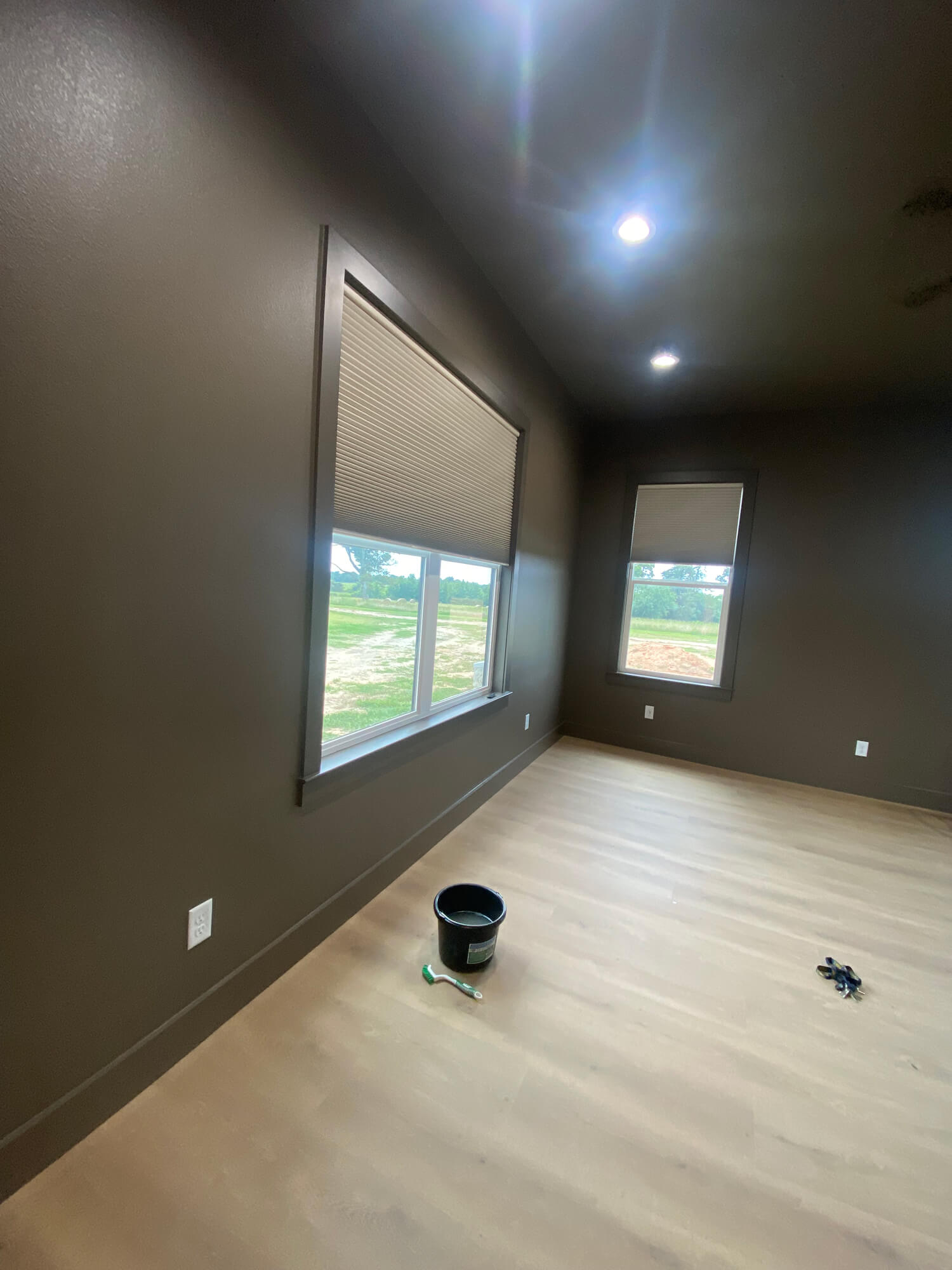 Texas Pro Painters: Recently painted room with dark brown walls, light wood-look flooring, two windows with blinds, and a paint bucket on the floor, showcasing interior painting in progress.