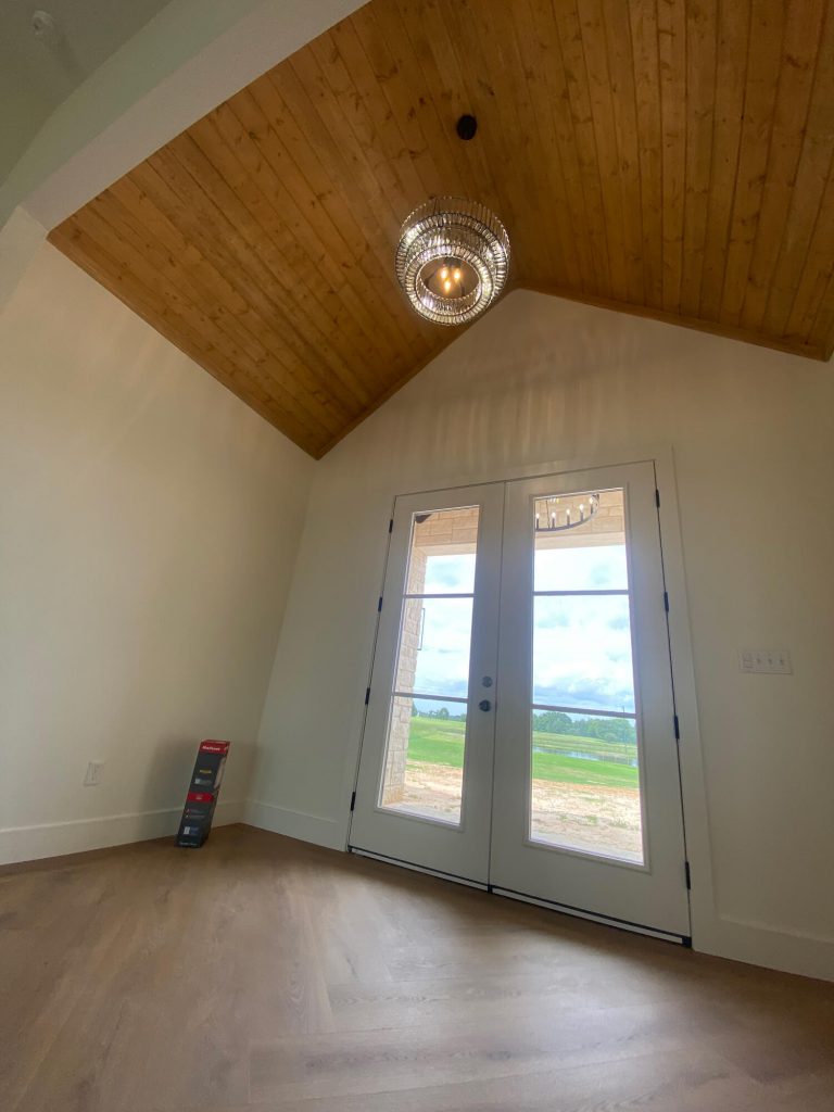Texas Pro Painters: Interior room with a vaulted wood-paneled ceiling, light beige walls, French doors overlooking a green landscape, and a modern chandelier, showcasing recent interior finishes.
