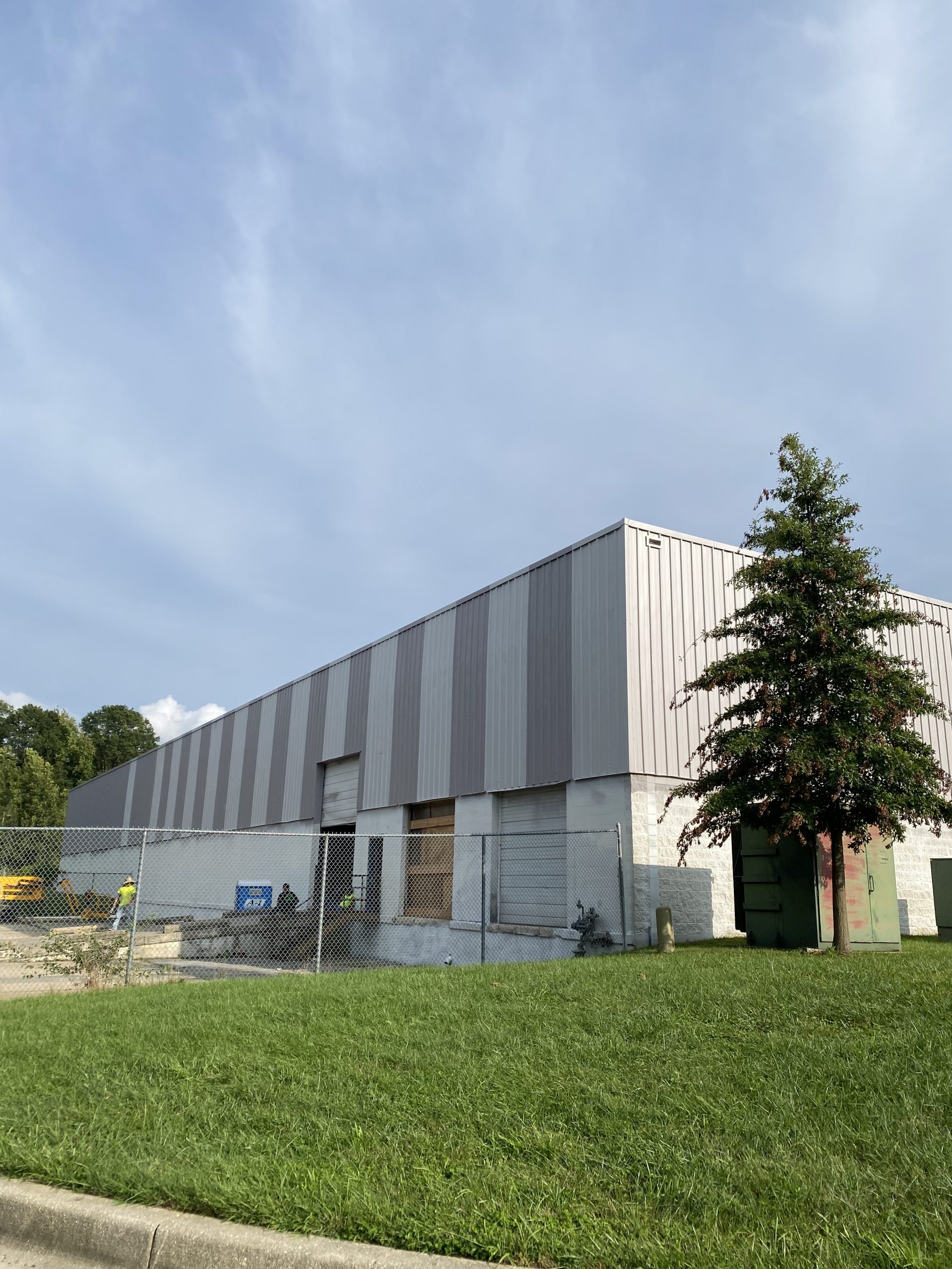 Commercial building exterior with vertical grey and white siding, a loading dock, a chain-link fence, and a mature evergreen tree in the foreground.