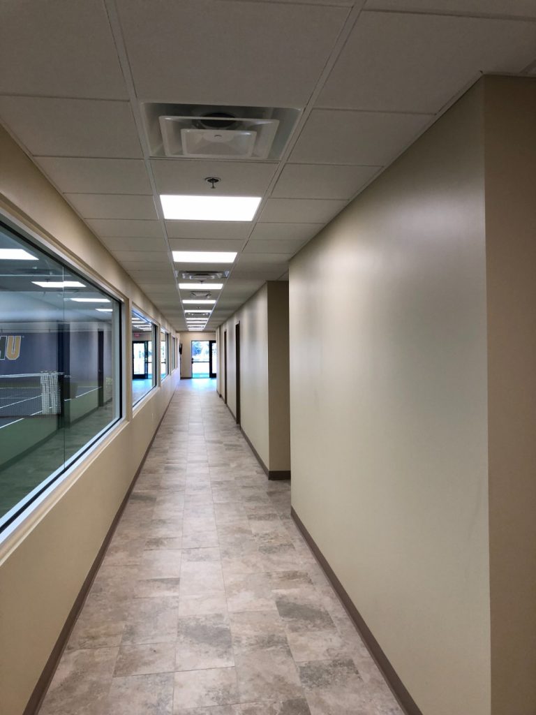 Long interior hallway with neutral-toned walls, tiled flooring, recessed lighting, and a large window looking into an indoor sports court.