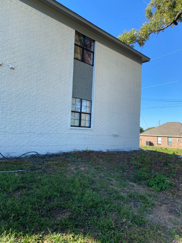 Exterior of a white brick building with two dark-framed windows vertically stacked, set against a clear blue sky and some green foliage.