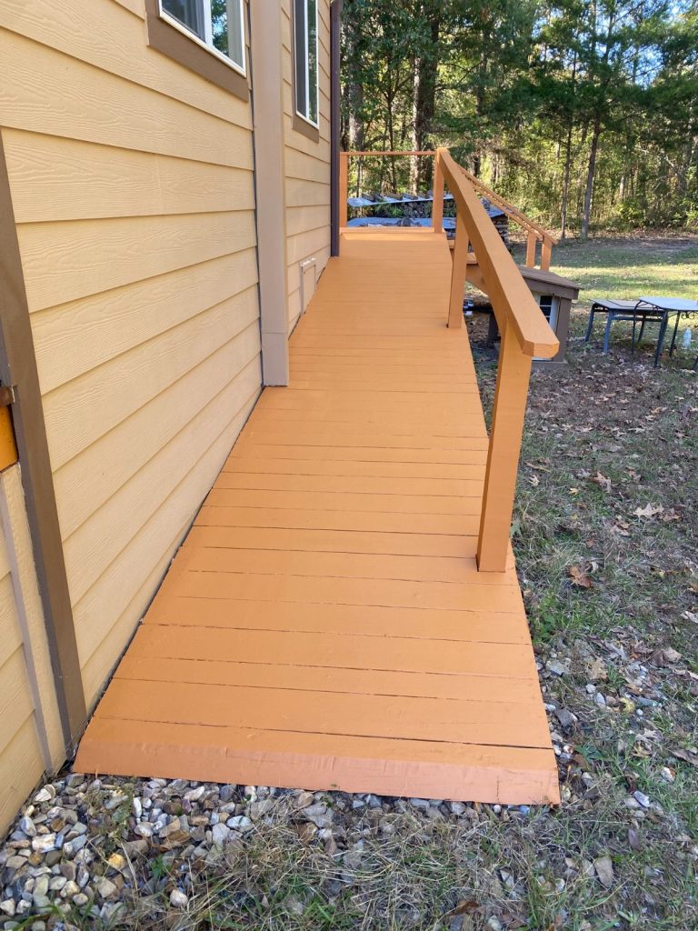Newly painted orange accessibility ramp leading to a tan-sided building, surrounded by green grass, gravel, and trees in the background.
