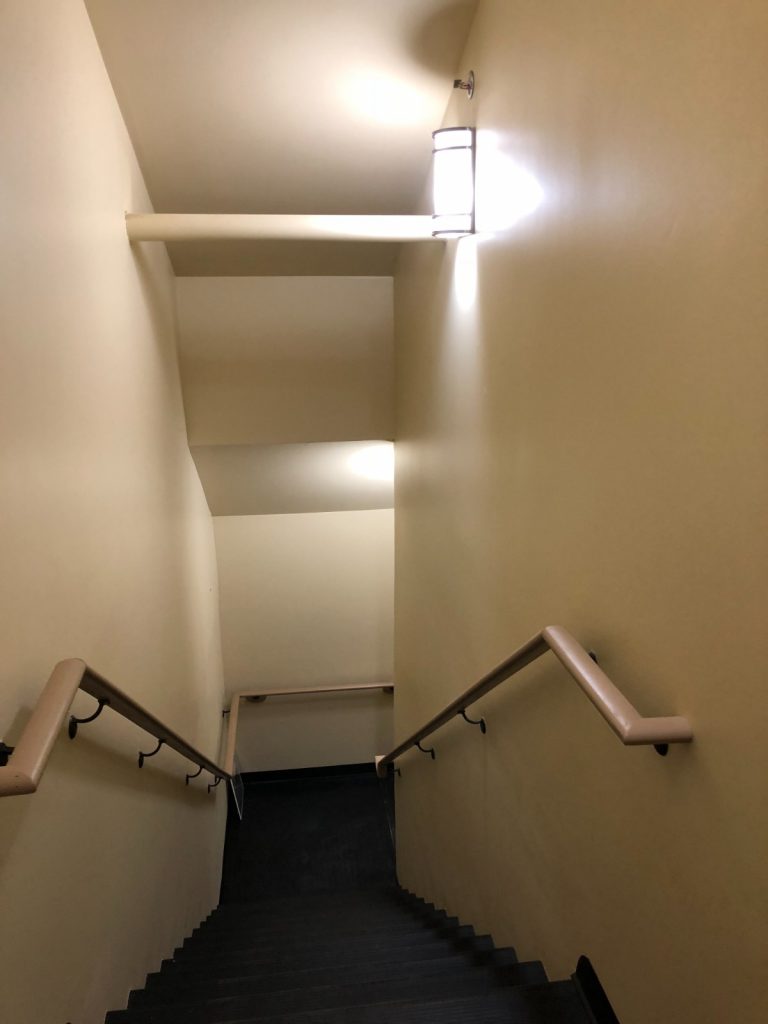 Interior staircase with light beige walls, black steps, light-colored handrails, and a modern wall sconce illuminating the landing.