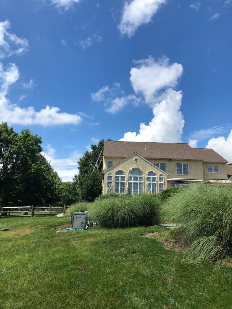 Exterior view of a large two-story house with yellow siding and a prominent sunroom, set amidst a lush green lawn and mature trees under a bright blue sky with white clouds.