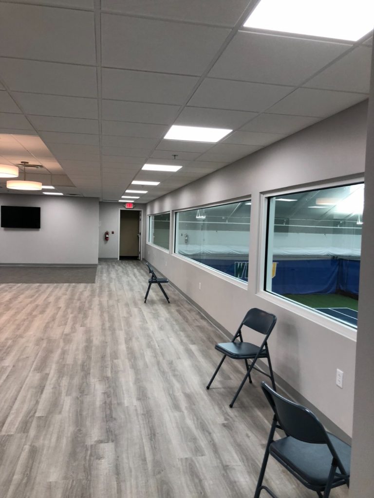 Modern commercial space featuring light grey walls, wood-look laminate flooring, windows overlooking an indoor tennis court, and black folding chairs.