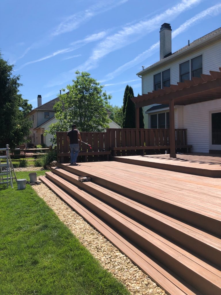 A worker applying stain to a large, multi-level wooden deck with a pergola, next to a white house and green lawn under a clear blue sky.