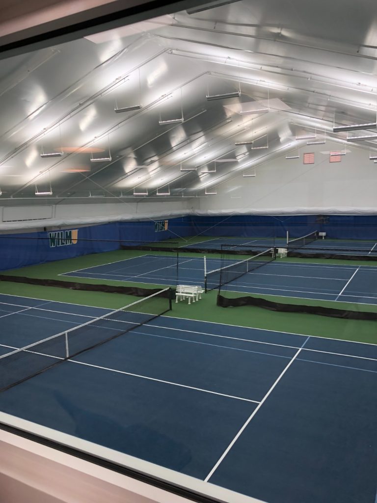 Indoor tennis facility with multiple blue and green courts, white nets, overhead lighting, and white benches, viewed from a slightly elevated perspective.