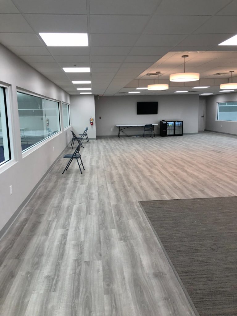 Large, modern commercial open space with light grey laminate flooring, white walls, multiple windows, a wall-mounted TV, and an area with carpet.