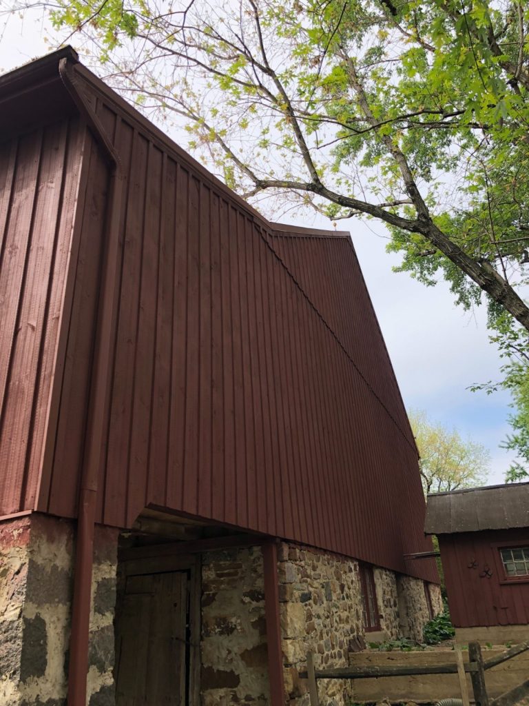 Large rustic barn with dark red vertical siding, a stone foundation, and a smaller red outbuilding, seen from below with tree branches overhead.