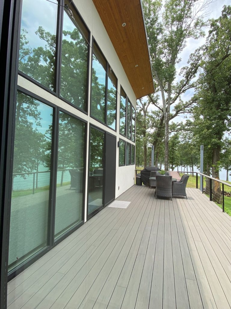 Modern lakeside home with large black-framed windows reflecting trees and the sky, a spacious outdoor deck with seating, and a view of the water.