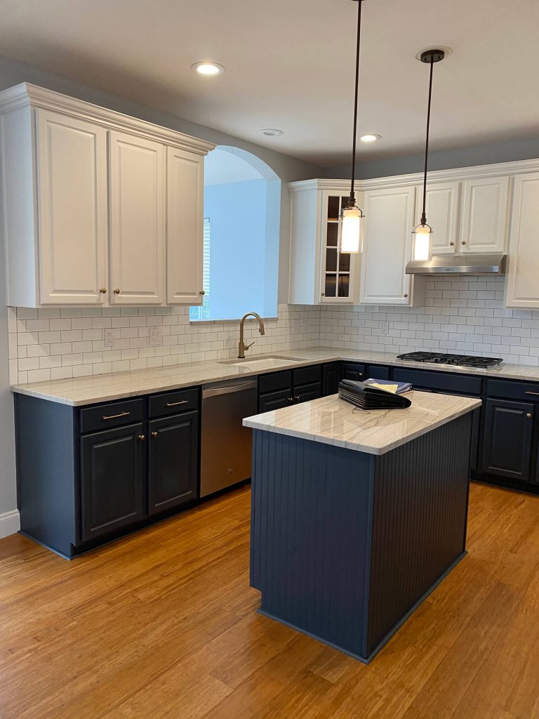 Texas Pro Painters: Newly renovated kitchen with two-toned cabinets (white uppers, dark grey lowers), a central island, subway tile backsplash, and light wood flooring, showcasing professional cabinet painting.